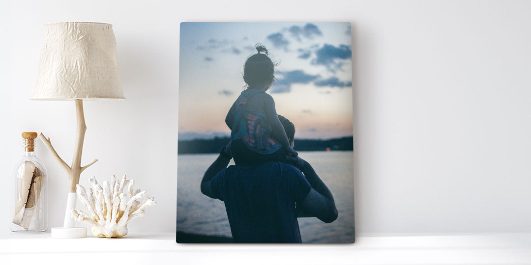 A metal wall art print of a man with a toddler on his shoulders overlooking the sunset rests besides beach summer decor.